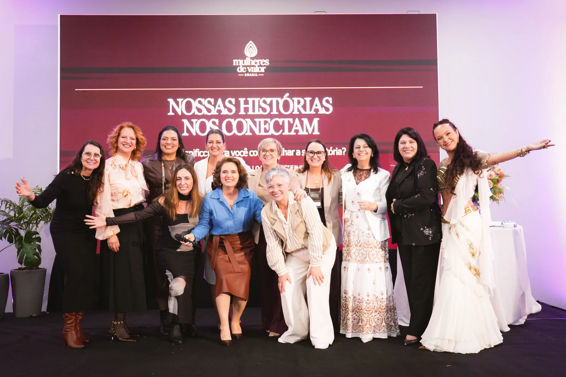 Mulheres reunidas no palco em frente ao painel Nossas histórias nos conectam, durante um encontro do Mulheres de Valor
